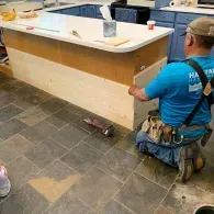 A worker in a blue shirt kneels on a tiled floor, installing light wood paneling onto the side of a kitchen island.
