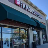The exterior of a Famous Footwear store with dark green awnings over large windows under a clear blue sky.