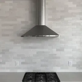 A stainless steel range hood centered above a black stovetop, set against a wall with light-gray, textured brick-style tiles.