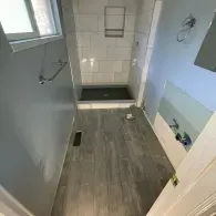 A partially renovated bathroom featuring gray wood-look tile flooring, light blue walls, and a white tiled shower stall.