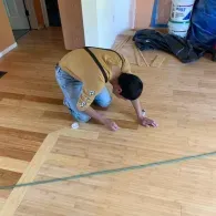 A person in a yellow shirt kneels on a partially installed hardwood floor, working on the planks in a residential room.