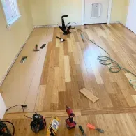 A room under renovation with partially installed light wood flooring, scattered power tools, and a coiled extension cord.