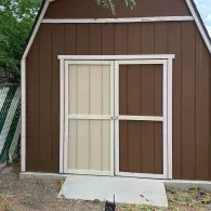 A brown wooden barn-style storage shed with white trim and mismatched light and dark brown double doors.
