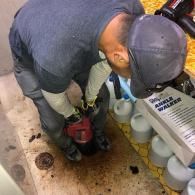 A person in a gray shirt and cap kneels on a floor to operate a red industrial cleaning machine near medical supplies.