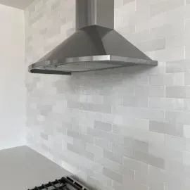A stainless steel range hood installed above a white, glossy subway tile backsplash in a kitchen.
