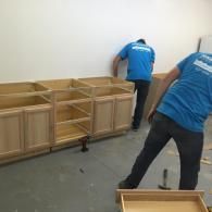 Two workers in matching blue shirts assemble wooden kitchen cabinet frames in a room with white walls and a gray floor.