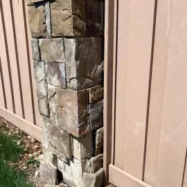 A stacked stone pillar stands between two beige vertical-panel fence sections, with a patch of grass at the base.