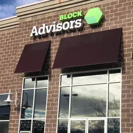 A brick storefront for Block Advisors with dark brown awnings over the windows and a green hexagon logo above the sign.