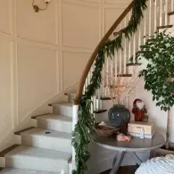 A curved staircase with a greenery garland on the railing, next to a table with a vase and decorative items.