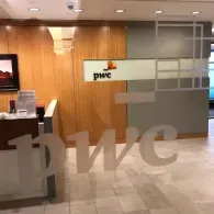 The lobby of a PwC office featuring light wood paneling, a gray wall, and the company logo on the wall and glass door.