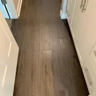 A hallway with dark wood-look plank flooring between a white door on the left and white cabinets on the right.