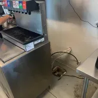 A soda fountain machine sits on a stainless steel counter in a commercial space, with exposed tubing connected to the wall.