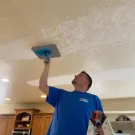 A person in a blue shirt uses a handheld trowel to smooth a textured ceiling in a kitchen.