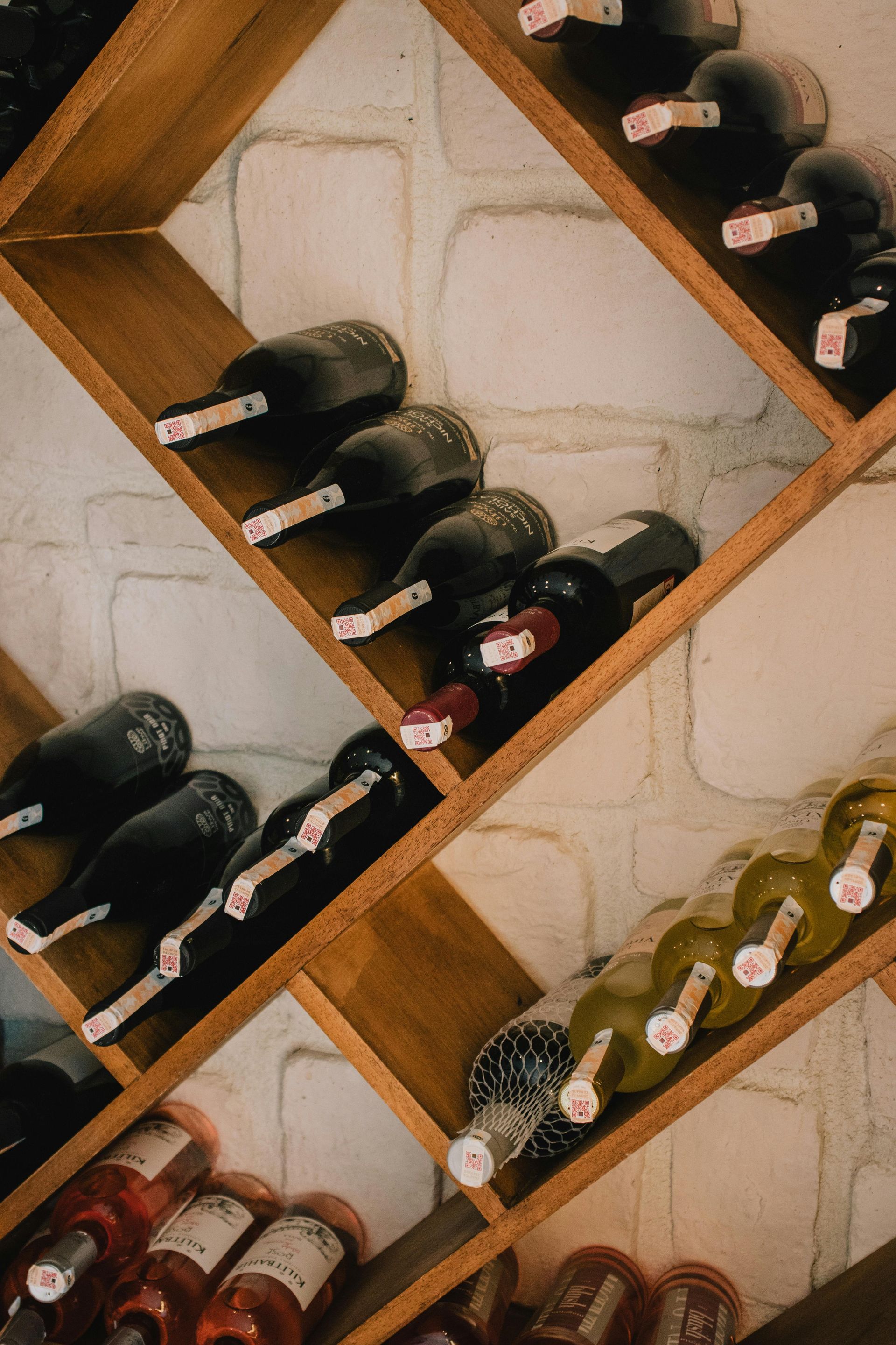 Wine bottles resting in wooden, diagonal cubbies against a white brick wall.