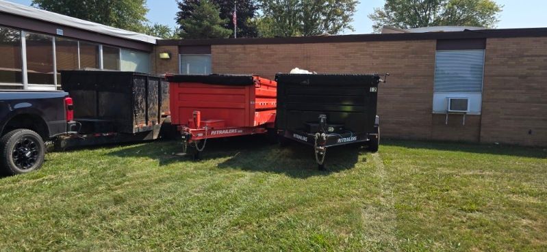 Three trailers parked on a grassy area next to a brick building. One is red, two are black.