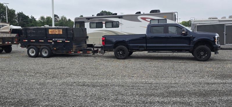 A dark blue truck pulling a trailer with a dumpster, parked on gravel.
