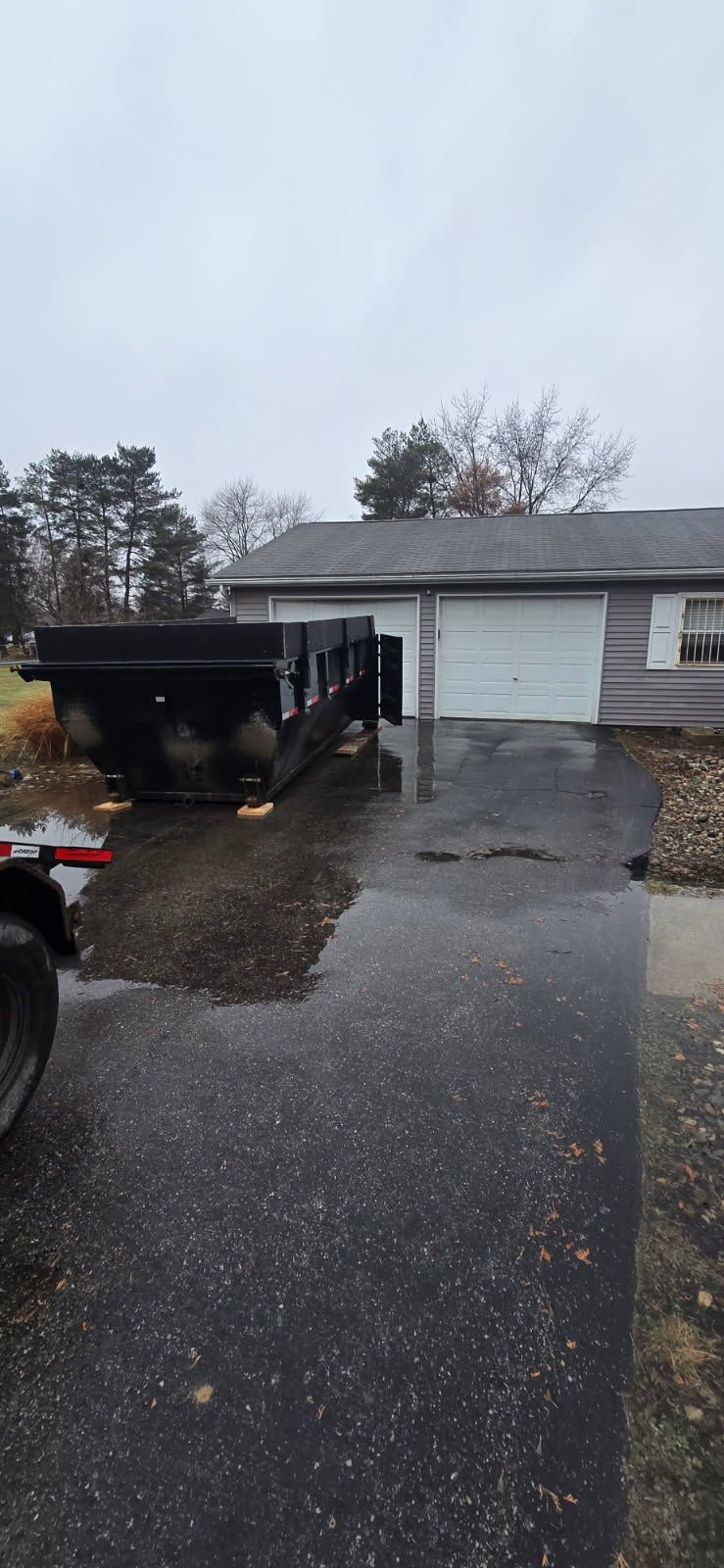 A black trailer is parked in front of a house on a wet asphalt driveway. Overcast sky.
