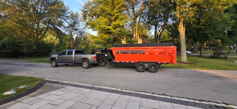 A gray pickup truck towing an orange dump trailer on a paved road with trees in the background.