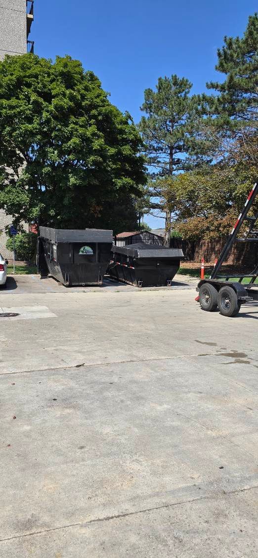 Black dumpsters on a concrete lot next to green trees, under a clear blue sky.