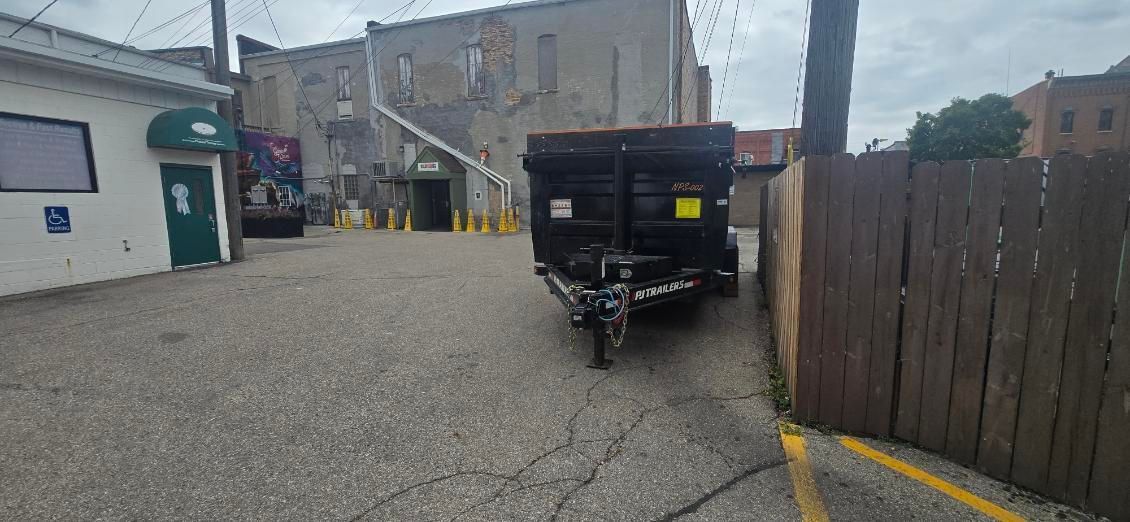 A black dumpster on a trailer parked in an alley, next to a wooden fence and a white building.