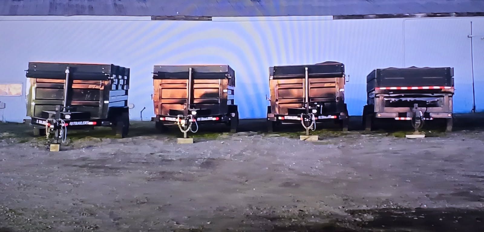 Four utility trailers parked in front of a white building on a gray surface.