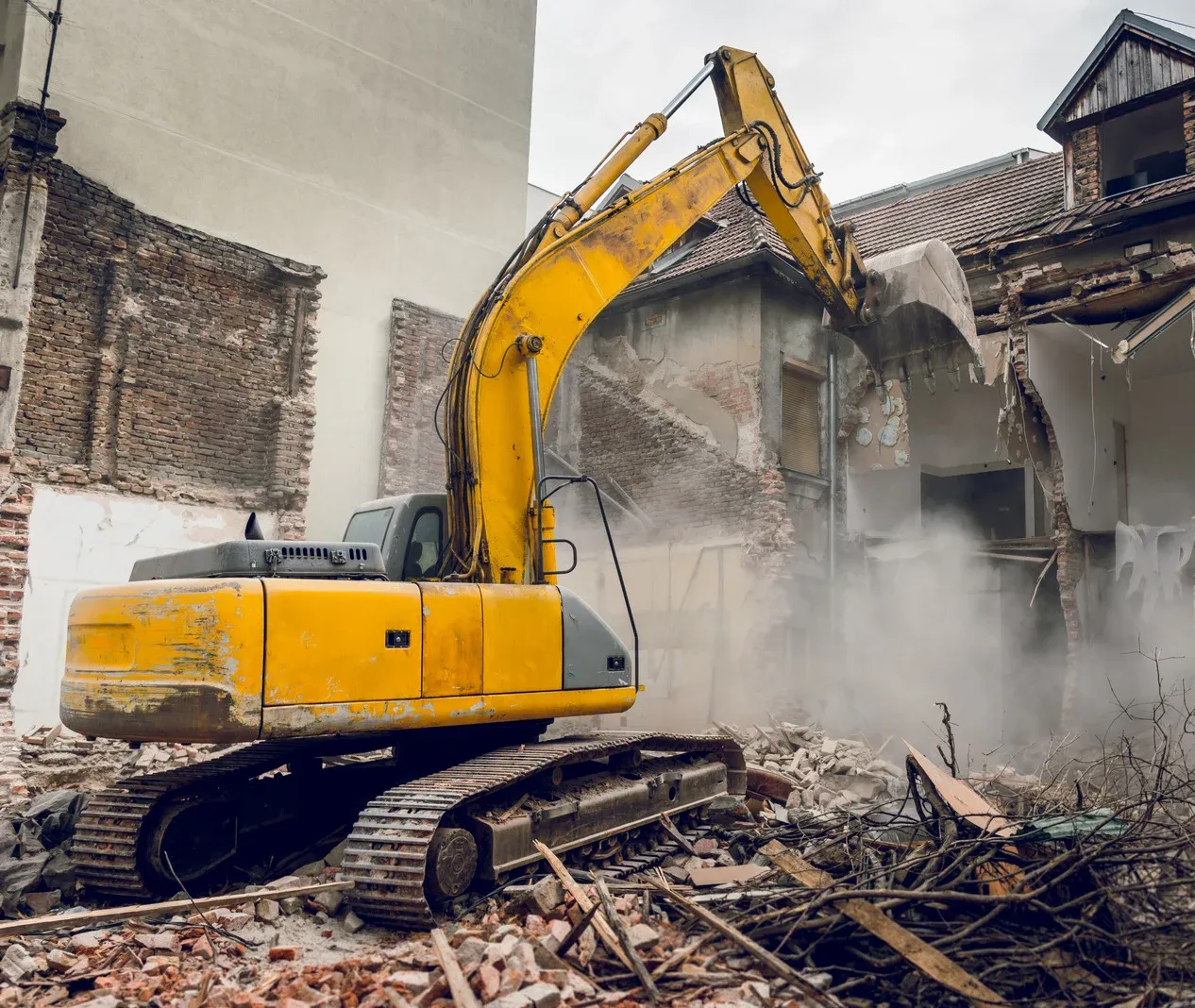 Yellow excavator demolishing a building, kicking up dust in a rubble-filled urban setting.
