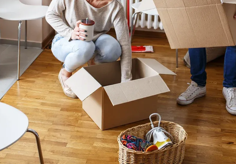 People packing moving boxes in a room with wooden floors. One person squats, another holds a box flap.