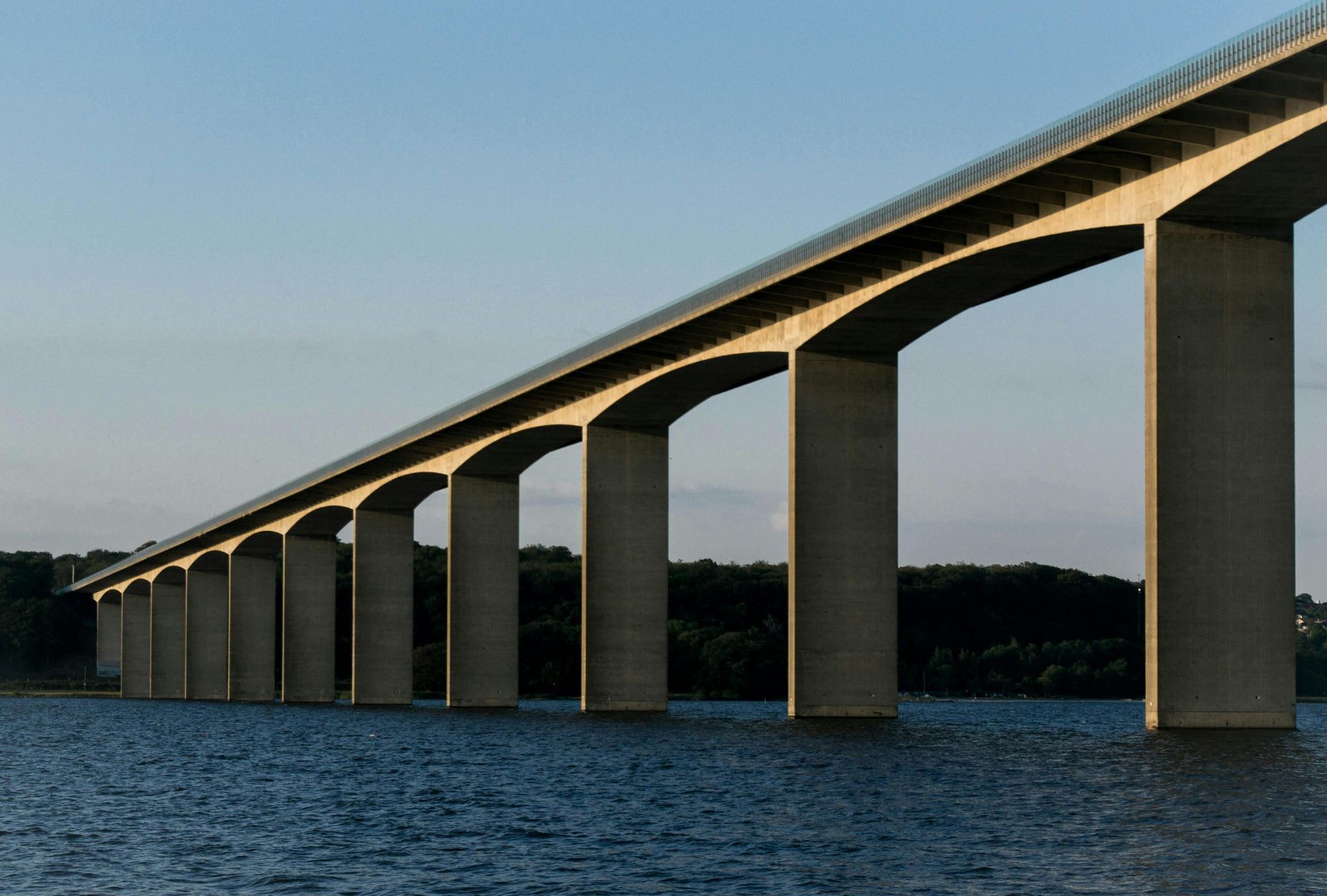 Concrete bridge spans over water, supported by pillars, under a light blue sky.