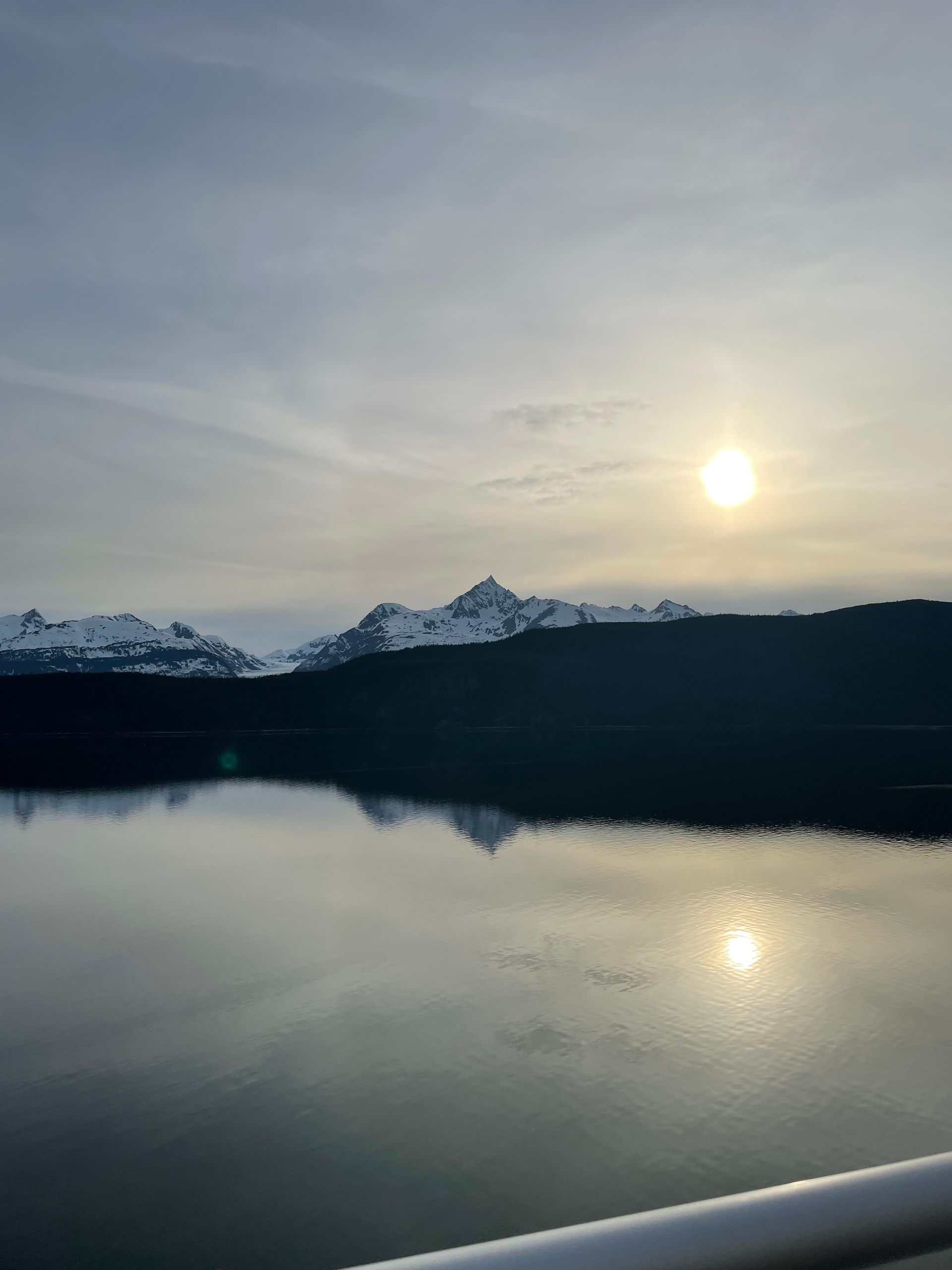 Calm waters in Alaska, snow capped mountains, and a bright sun behind a thin veil of clouds. 