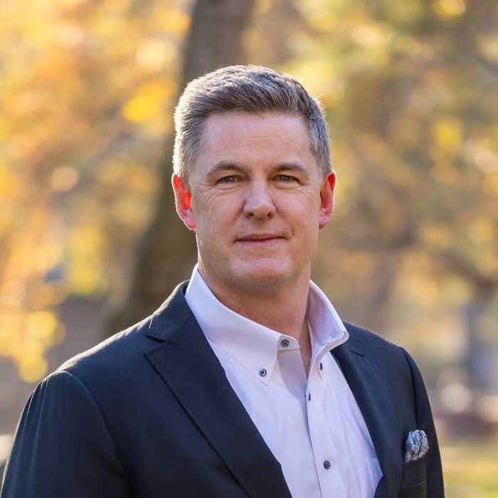 A woman wearing a black jacket and a necklace smiles for the camera