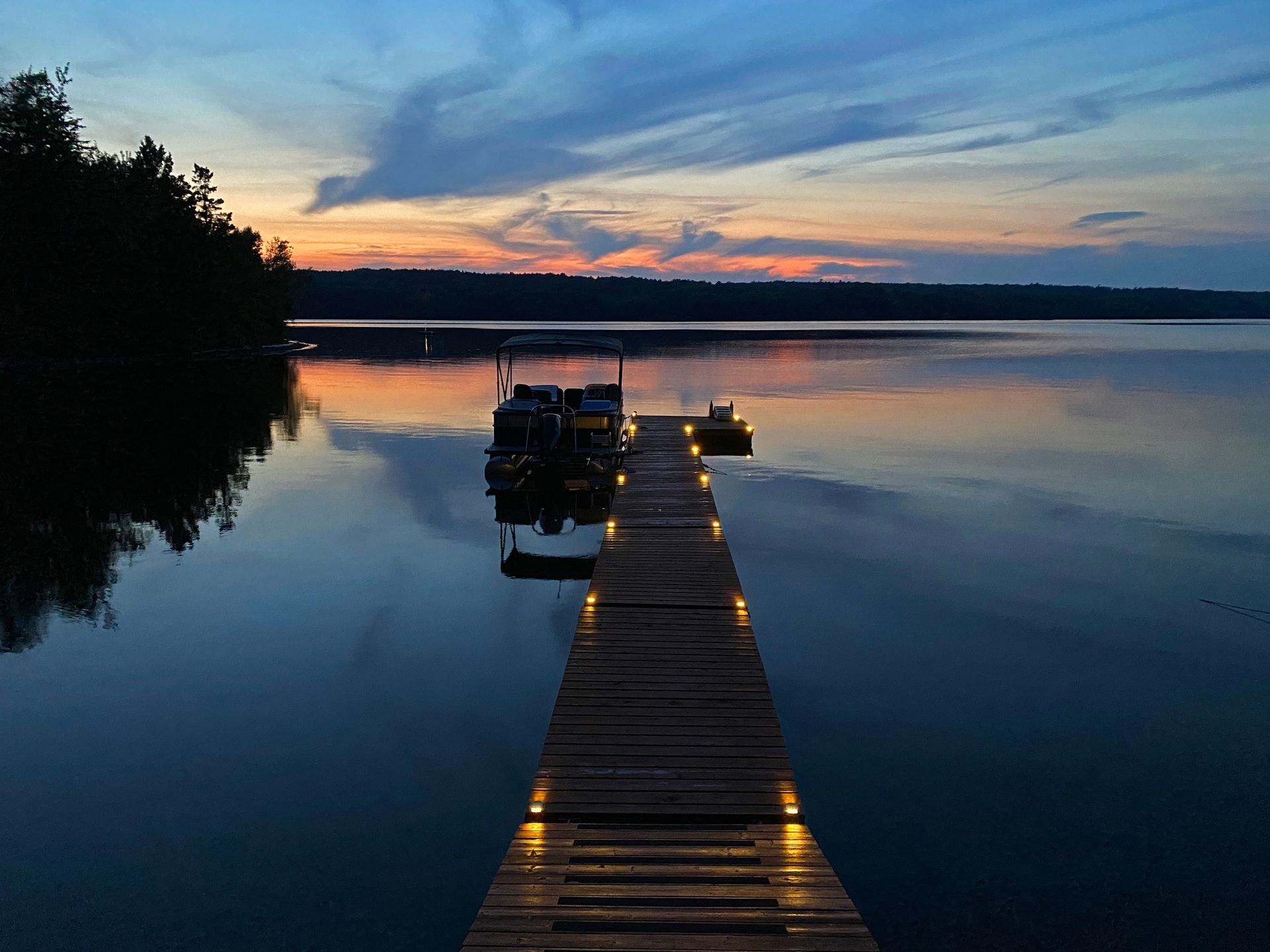 A wooden dock with lights on it leading to a lake at sunset.