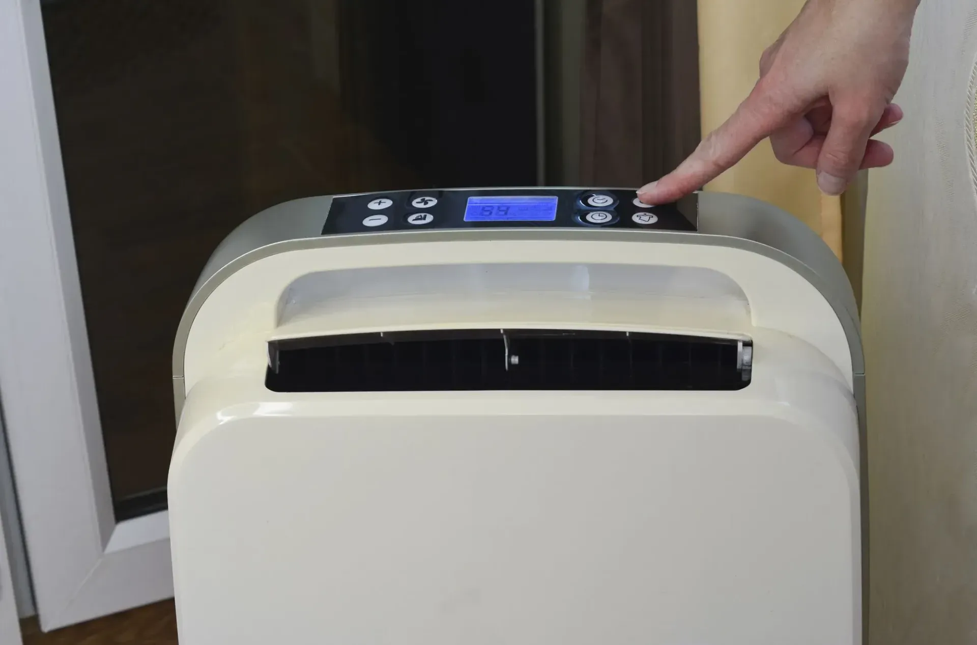 A person’s finger presses a button on the control panel of a cream-colored portable dehumidifier.