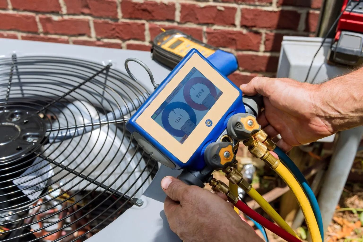 A person uses a digital HVAC manifold gauge to check refrigerant levels on an outdoor air conditioning unit.