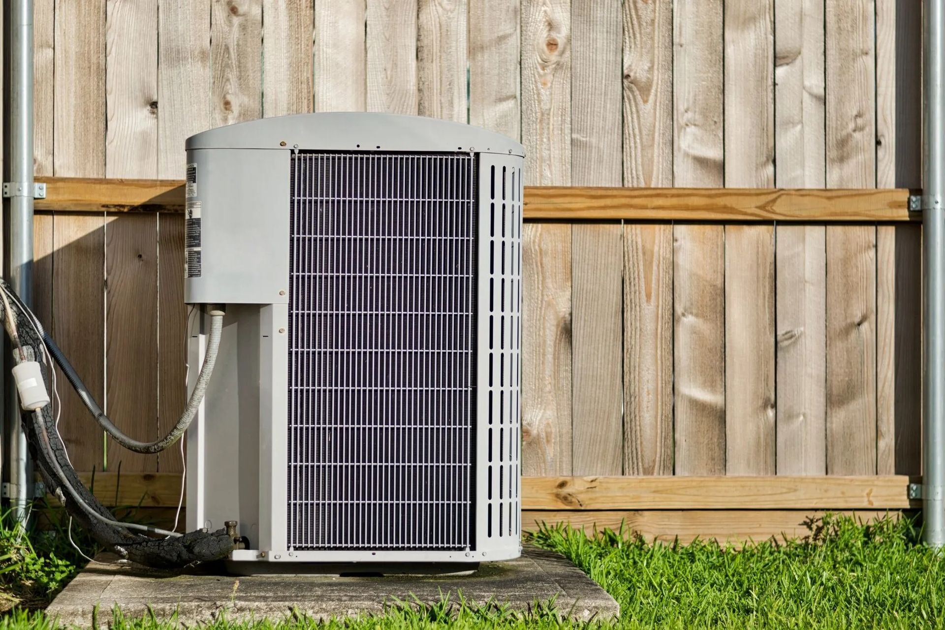 A gray air conditioner unit stands on a concrete pad in front of a wooden fence, surrounded by green grass.