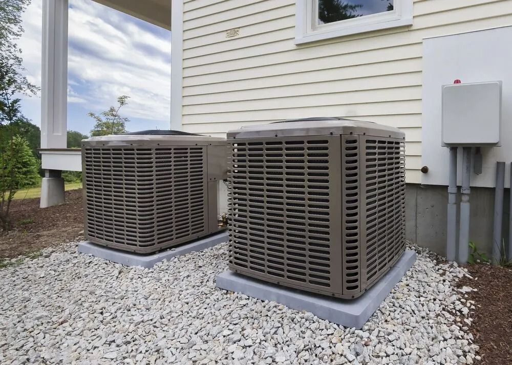 Two tan outdoor HVAC units sit on gravel pads next to the beige siding of a house.