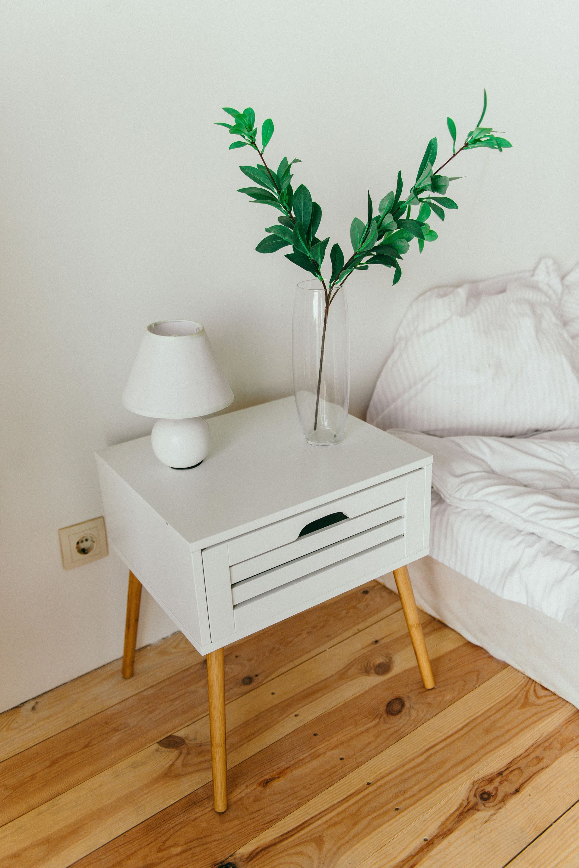 A white nightstand with a lamp and a vase of leaves on it in a bedroom.