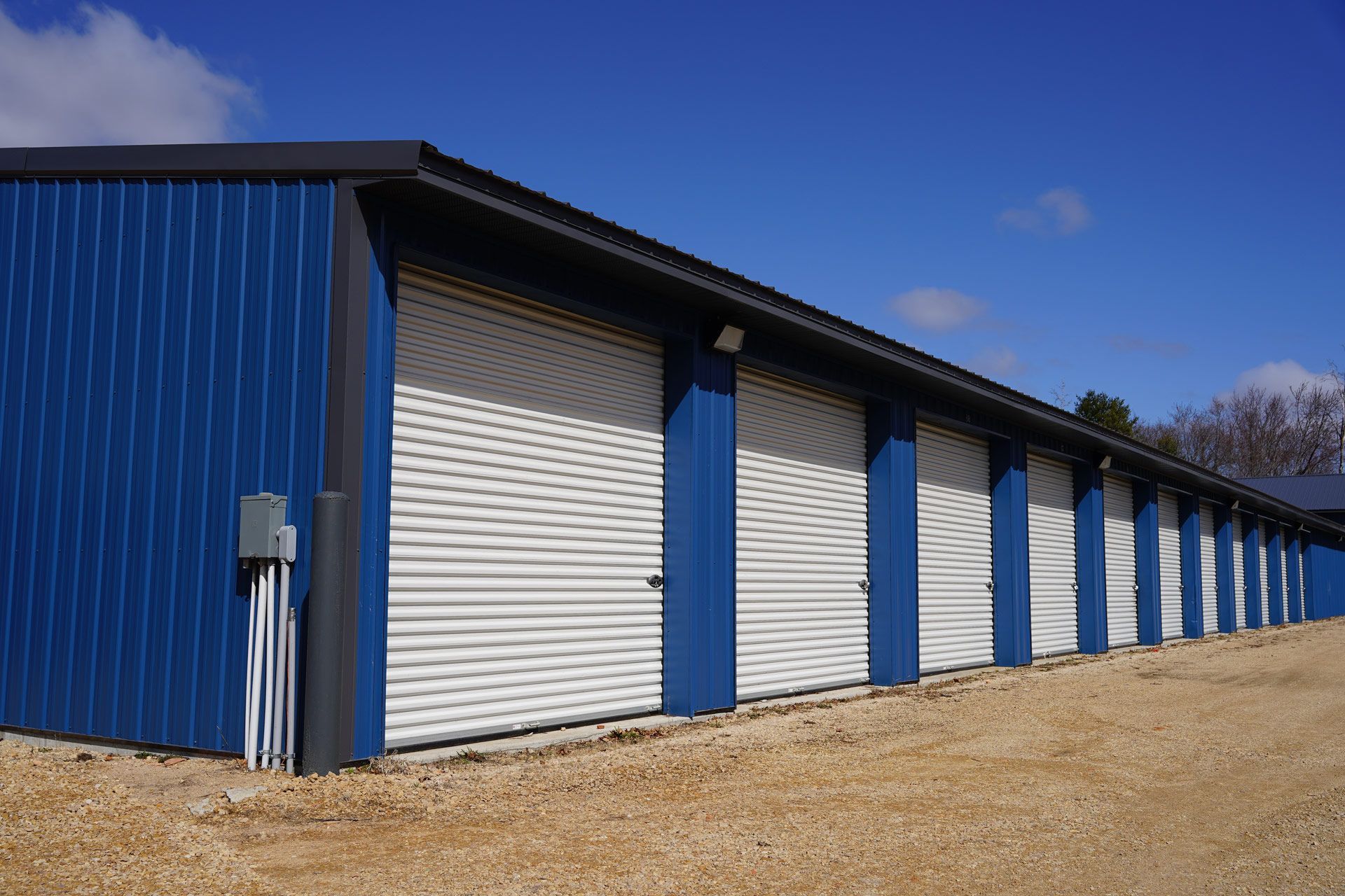 Blue storage units with white roll-up doors, on a gravel lot under a blue sky.