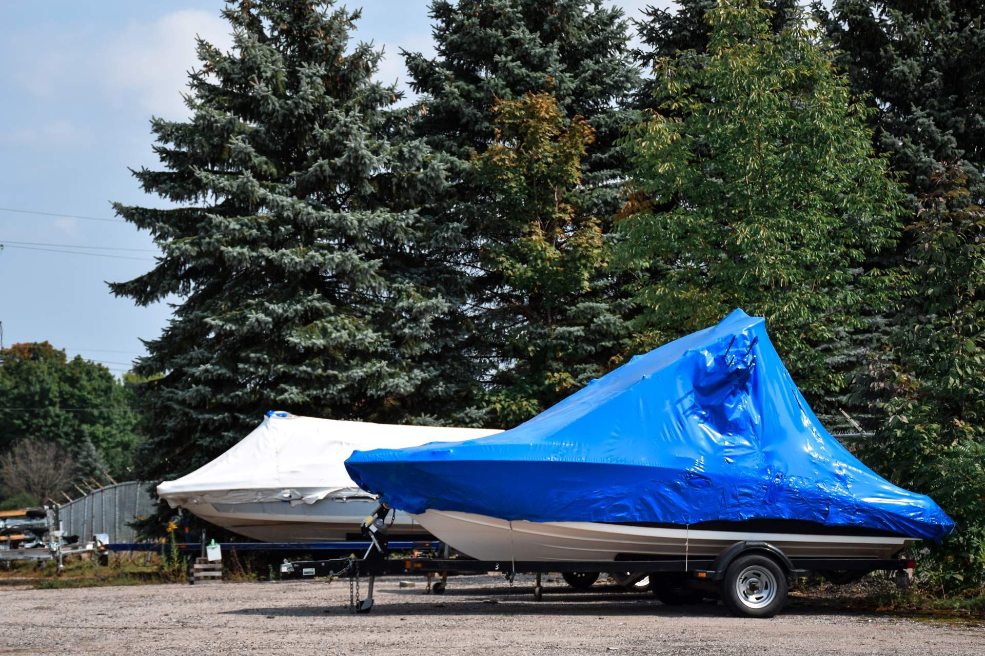 Boats covered in blue and white tarps parked on trailers, trees in the background.