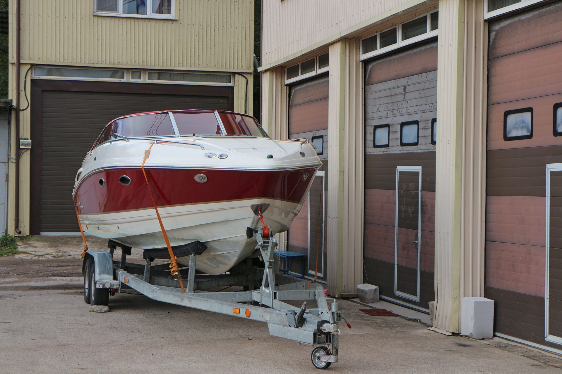 A red and white boat on a trailer parked next to a garage with brown doors.