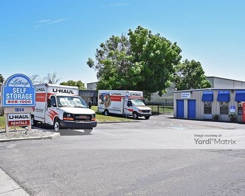 U-Haul self-storage facility with two trucks parked outside. Blue sign, buildings, and a tree are visible.