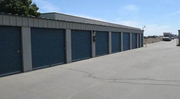 Storage units with blue doors in a row, with a gray roof and concrete ground, under a blue sky.