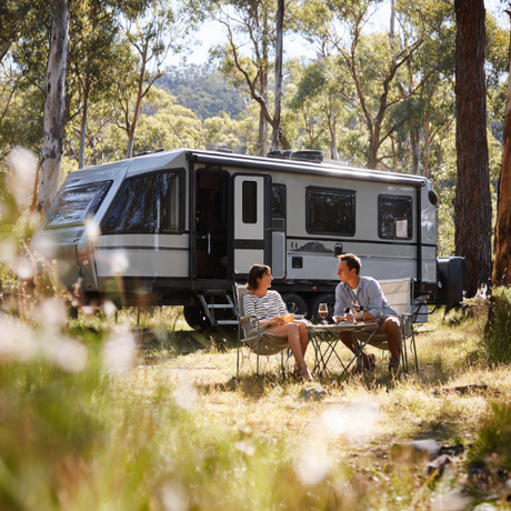 Couple sitting at table in front of a gray travel trailer in a wooded area.
