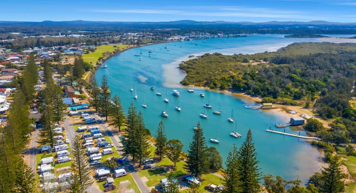Aerial view of Yamba campground with cabins and RVs beside a waterway. Blue sky, water, and green trees.