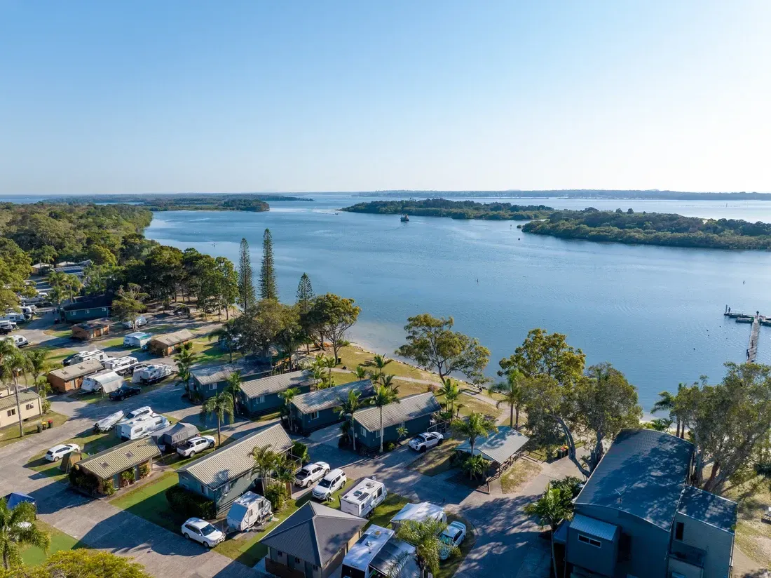 Aerial view of Yamba campground with cabins and RVs beside a waterway. Blue sky, water, and green trees.