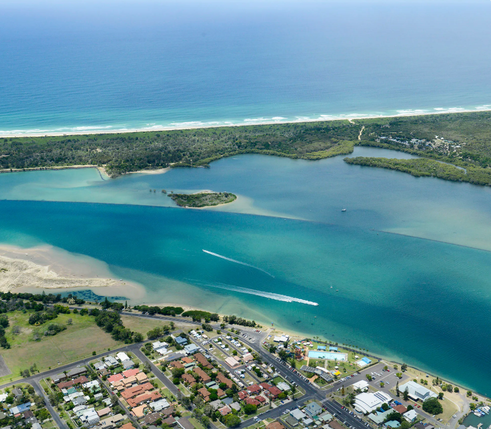Aerial view of Yamba campground with cabins and RVs beside a waterway. Blue sky, water, and green trees.
