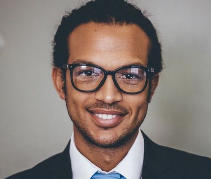 Man in glasses smiles, wearing a suit and tie, against a neutral background.