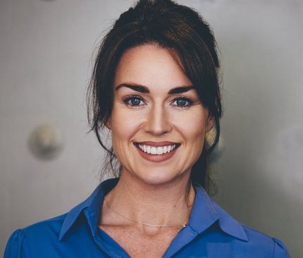Woman with dark hair, wearing a blue shirt, smiling, looking at the camera. Neutral background.