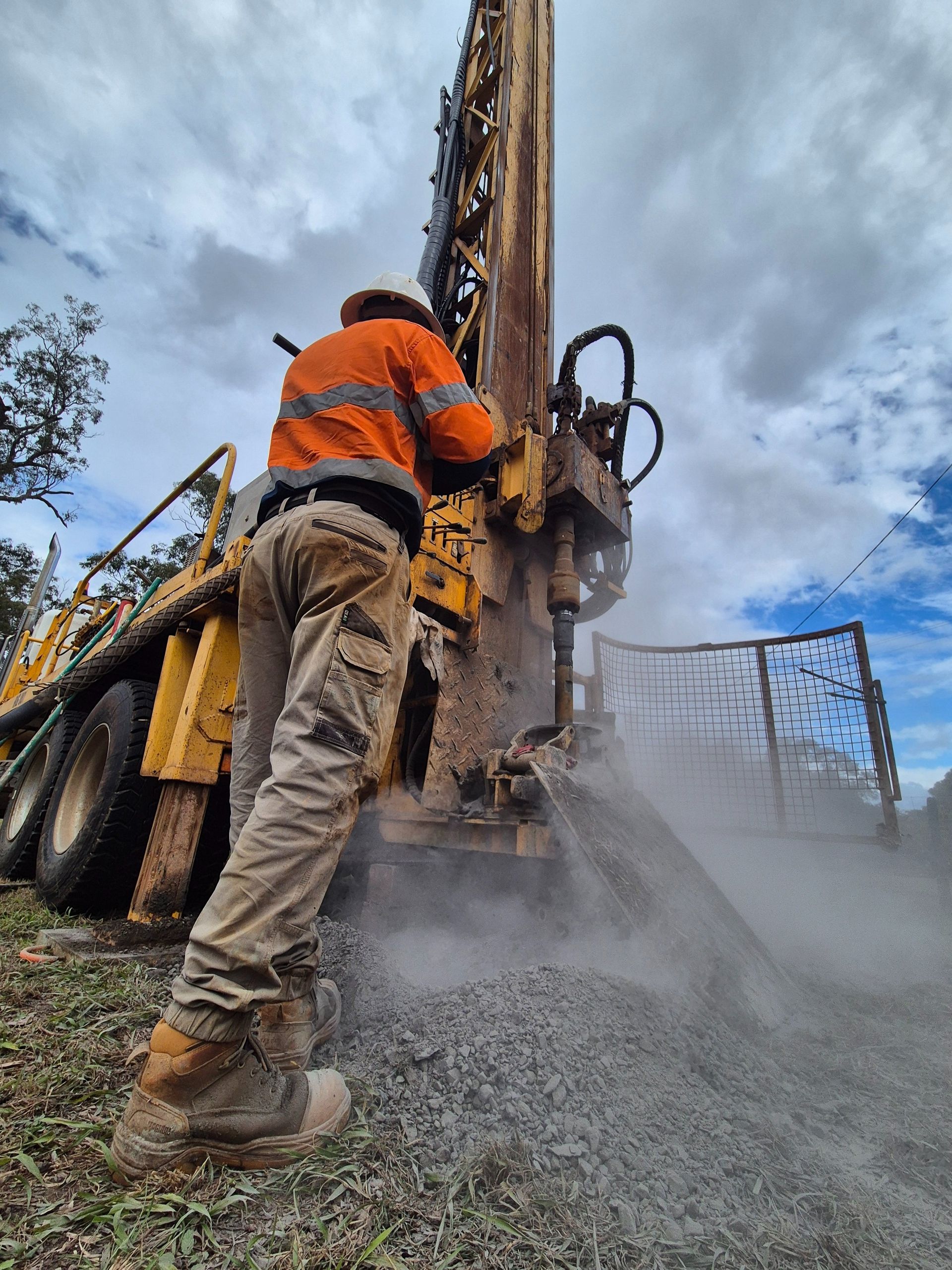 Man in orange workwear operating drilling equipment outdoors. Dust and debris present — Local Drilling Services In Kingaroy, QLD