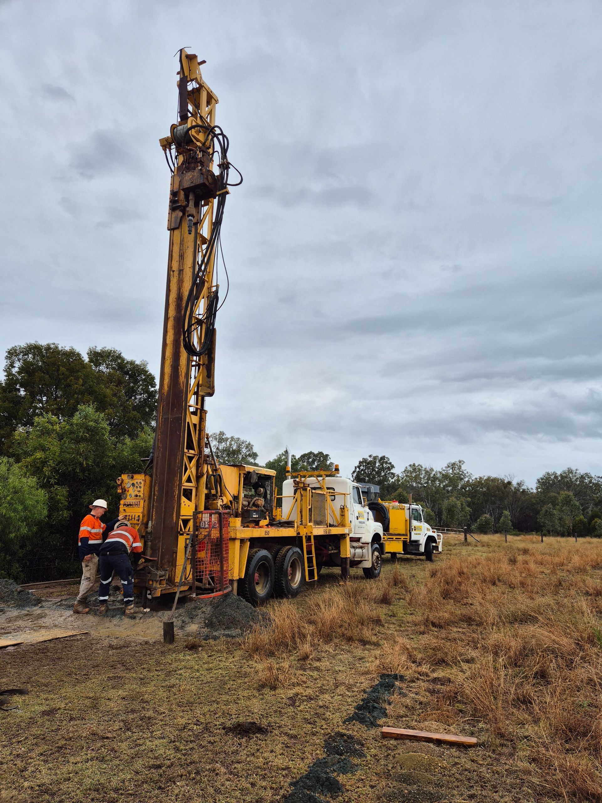 Yellow drilling rig in field with two workers — Local Drilling Services In North Burnett, QLD