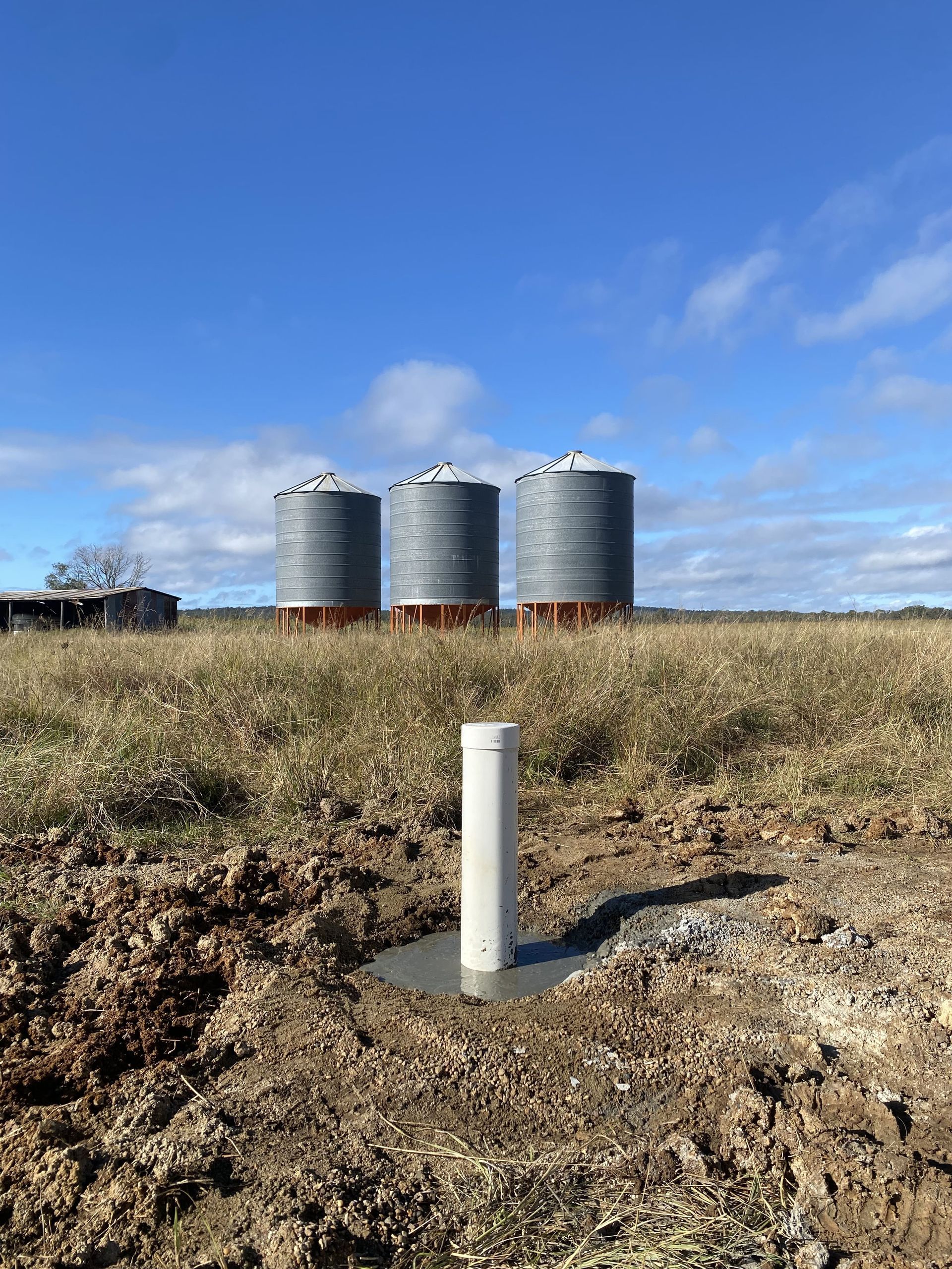A white PVC pipe sticking out of a concrete pad, in a field with three metal silos in the background — Local Drilling Services In Kingaroy, QLD